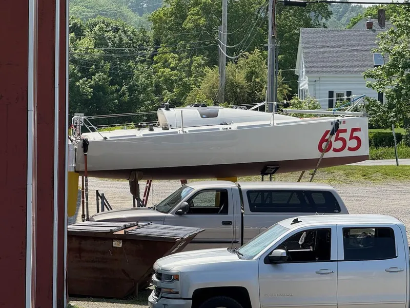 Slide: The Image of 2006 Mas Mini Transat 6.50 sailboat on trailer, parked near trucks, surrounded by greenery. - 22