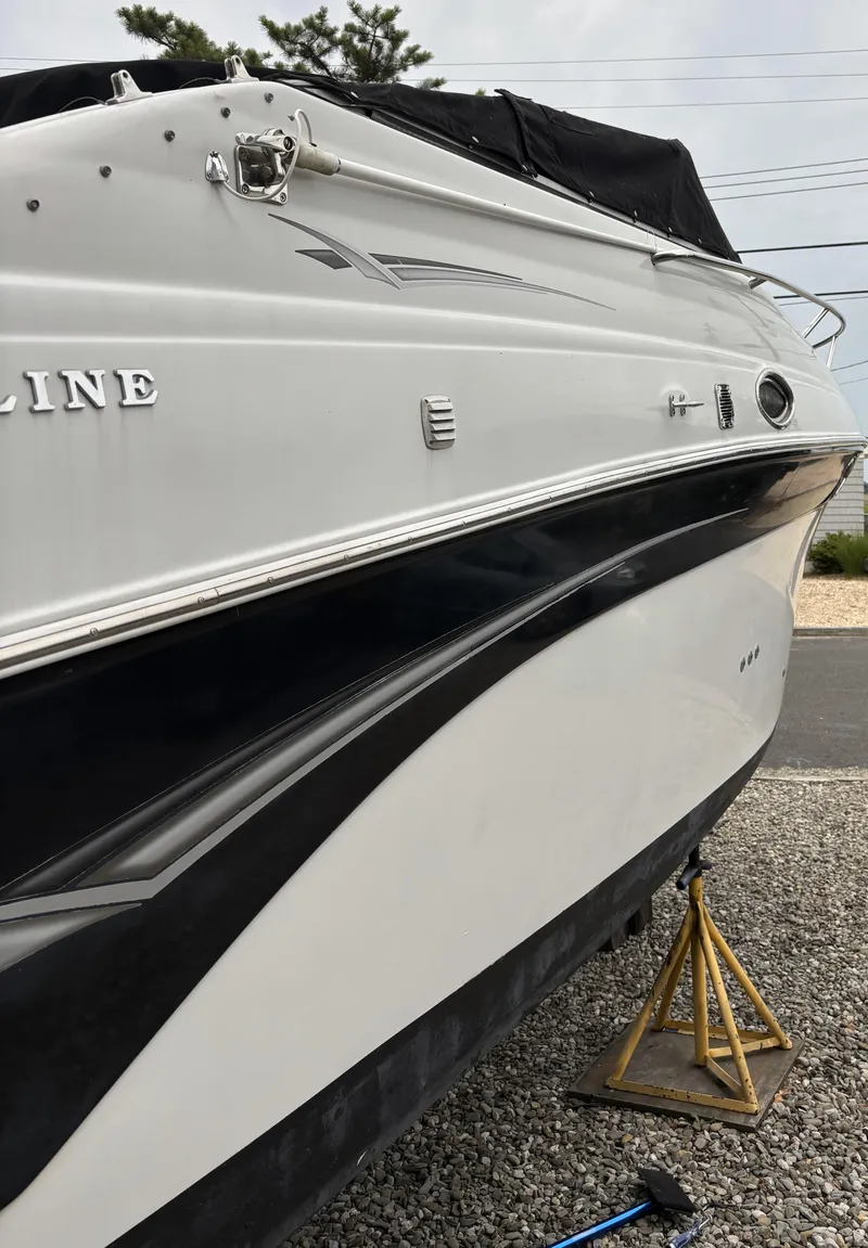 Slide: The Image of 2003 Crownline 262 CR boat on stands, white and black hull, parked on gravel. - 11