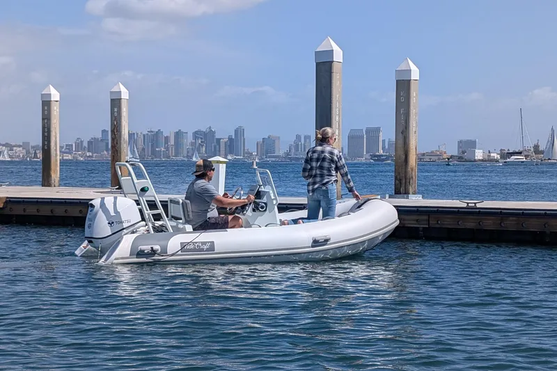 Slide: The Image of Two people on a 2025 Tide Craft Nantucket 12 boat near a dock, city skyline in background. - 2