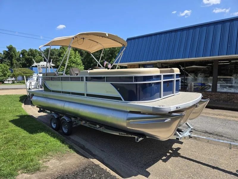 Slide: The Image of 2026 Godfrey Xperience 2286 SBX pontoon boat on trailer, parked near a blue-roofed building. - 3