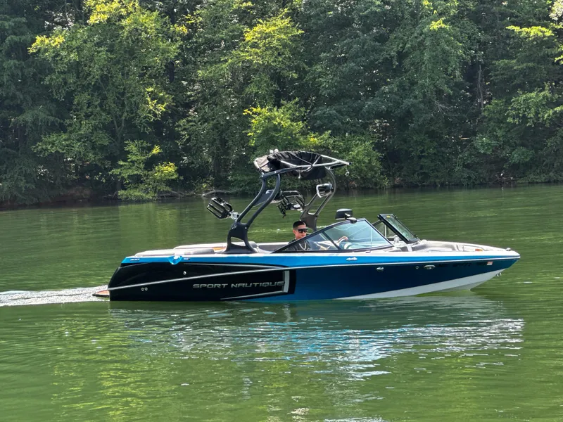 Slide: The Image of 2014 Nautique Sport 200 boat cruising on a serene lake with lush green trees. - 2