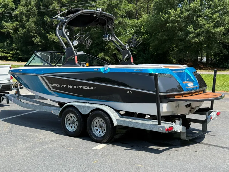 Slide: The Image of 2014 Nautique Sport 200 boat on trailer, parked outdoors, surrounded by trees. - 25