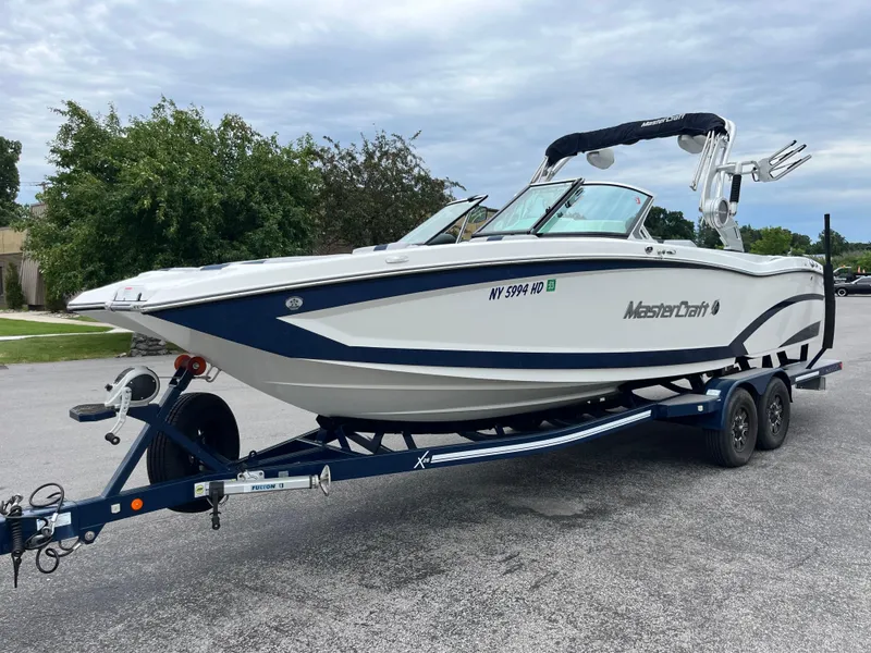 Slide: The Image of 2019 MasterCraft X26 boat on trailer, parked outdoors, with trees in the background. - 31