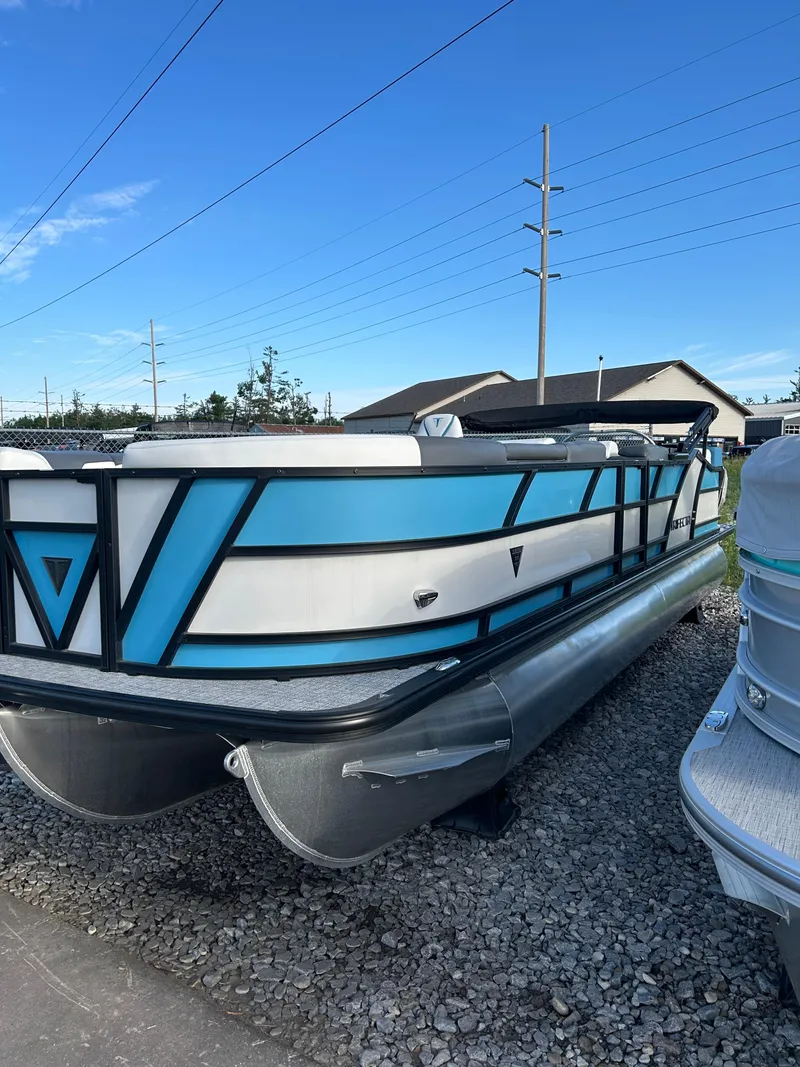 Slide: The Image of 2024 Trifecta 25C SS pontoon boat with blue and white design on gravel lot. - 1
