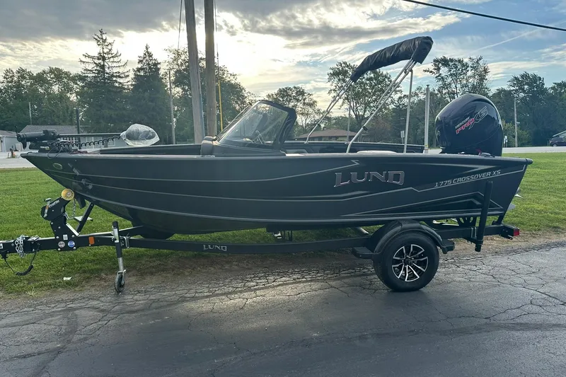 Slide: The Image of 2026 Lund 1775 Crossover XS boat on trailer, parked outdoors, with trees in background. - 36