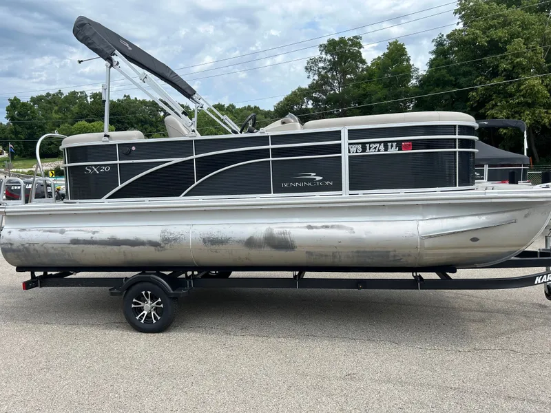 The Image of A 2019 Bennington 20SSRCX pontoon boat on a trailer, featuring a black canopy and silver hull. - 1