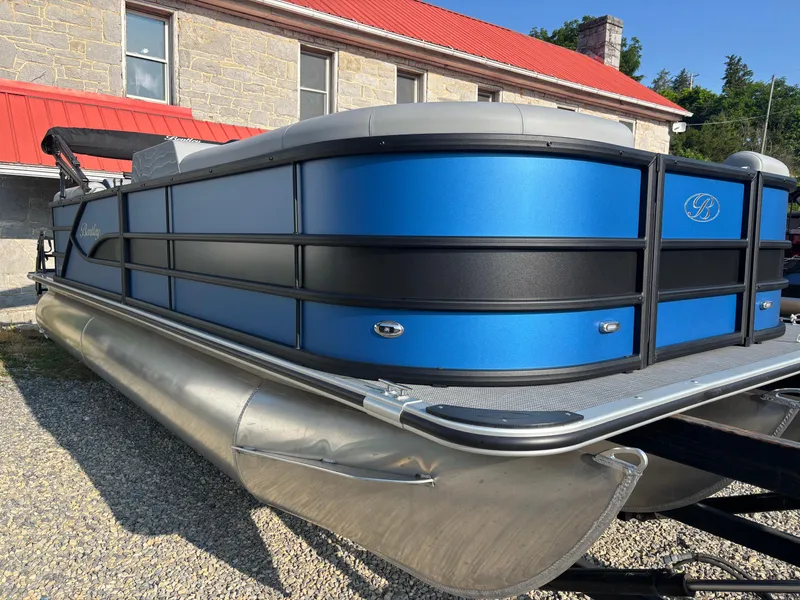 Slide: The Image of 2025 Bentley Pontoons Legacy Navigator, blue and black design, parked on gravel near a building. - 2