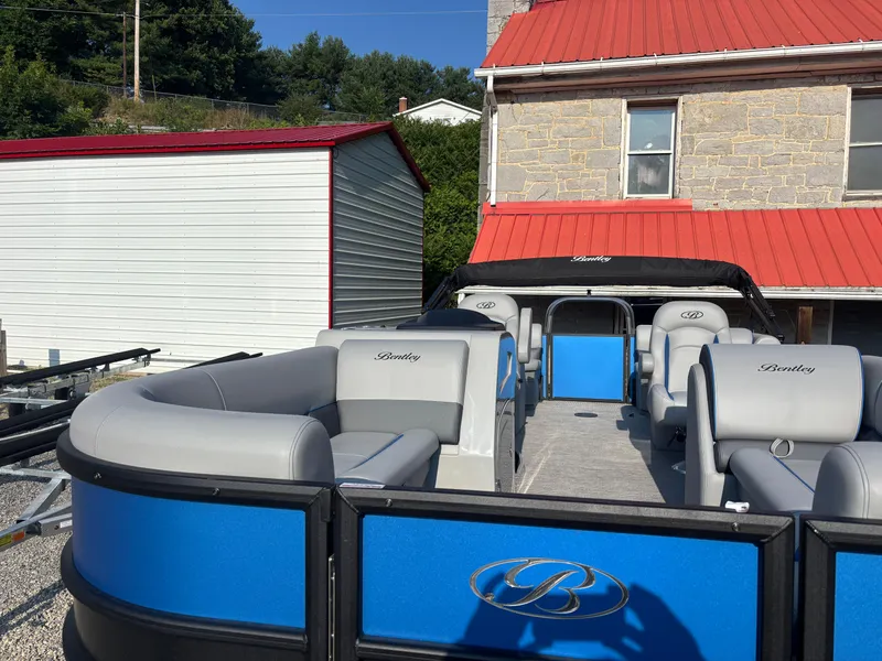 Slide: The Image of 2025 Bentley Pontoons Legacy Navigator with blue and gray seating, parked near a red-roofed building. - 8