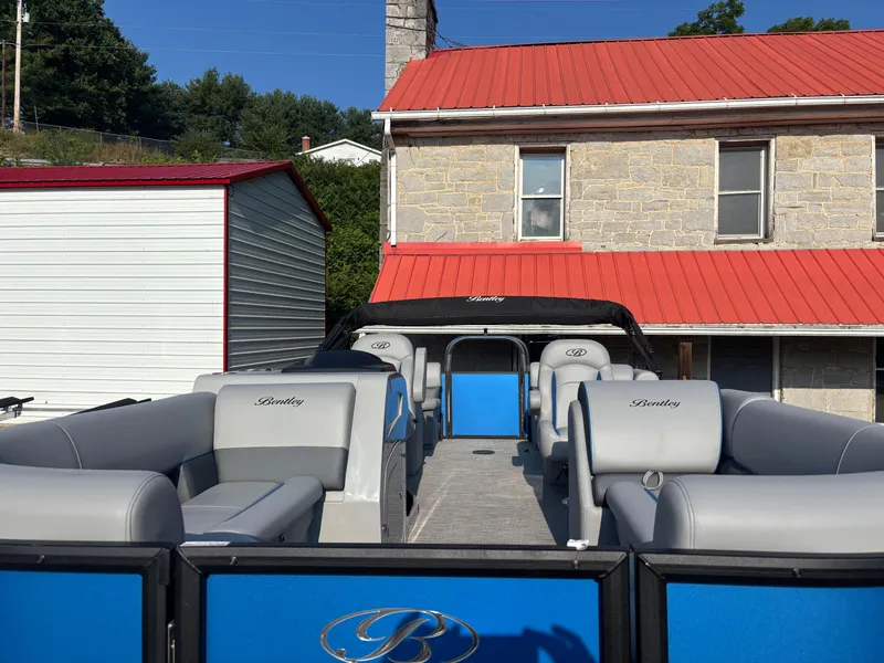 Slide: The Image of 2025 Bentley Pontoons Legacy Navigator with gray seating, parked near a red-roofed building. - 9