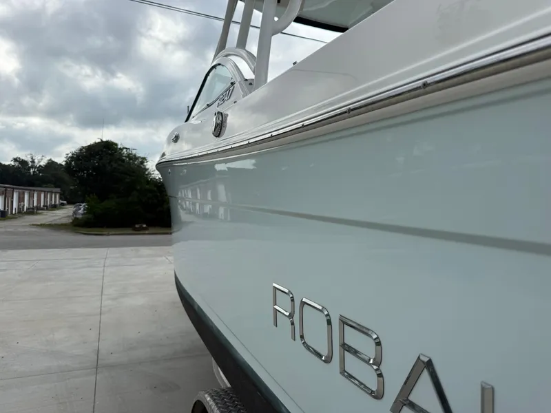 Slide: The Image of 2018 Robalo R247 Dual Console boat side view with logo, parked outdoors. - 6