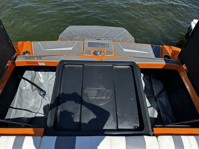Slide: The Image of 2023 Malibu 21 LX boat interior with orange accents and storage compartment, near water. - 22