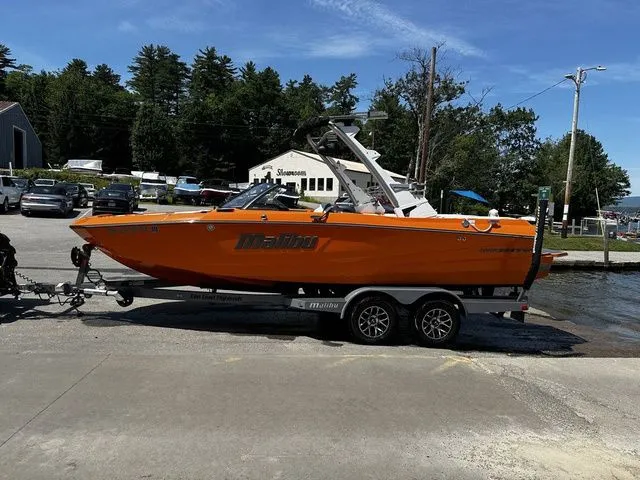 Slide: The Image of 2023 Malibu 21 LX boat in vibrant orange on a trailer at a lakeside dock. - 25