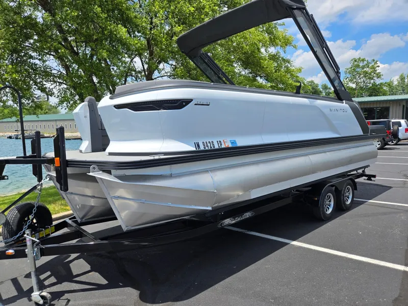 Slide: The Image of 2024 Manitou Cruise pontoon boat on trailer, parked near a lake with trees in the background. - 7