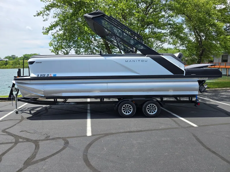 Slide: The Image of 2024 Manitou Cruise pontoon boat on trailer, parked near a lake. - 8