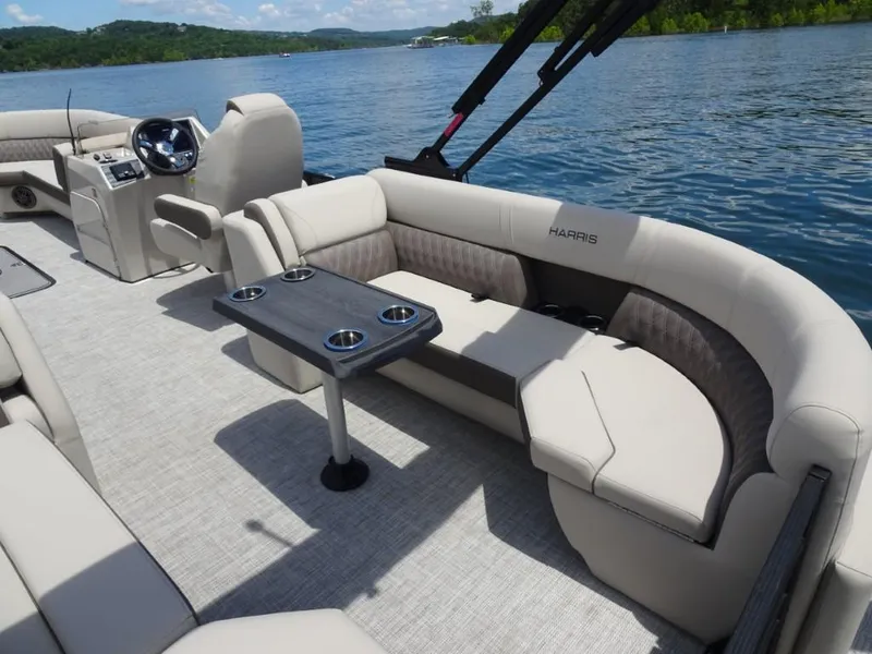 Slide: The Image of 2023 Harris Sunliner 250 pontoon boat interior with plush seating and table on a scenic lake. - 11