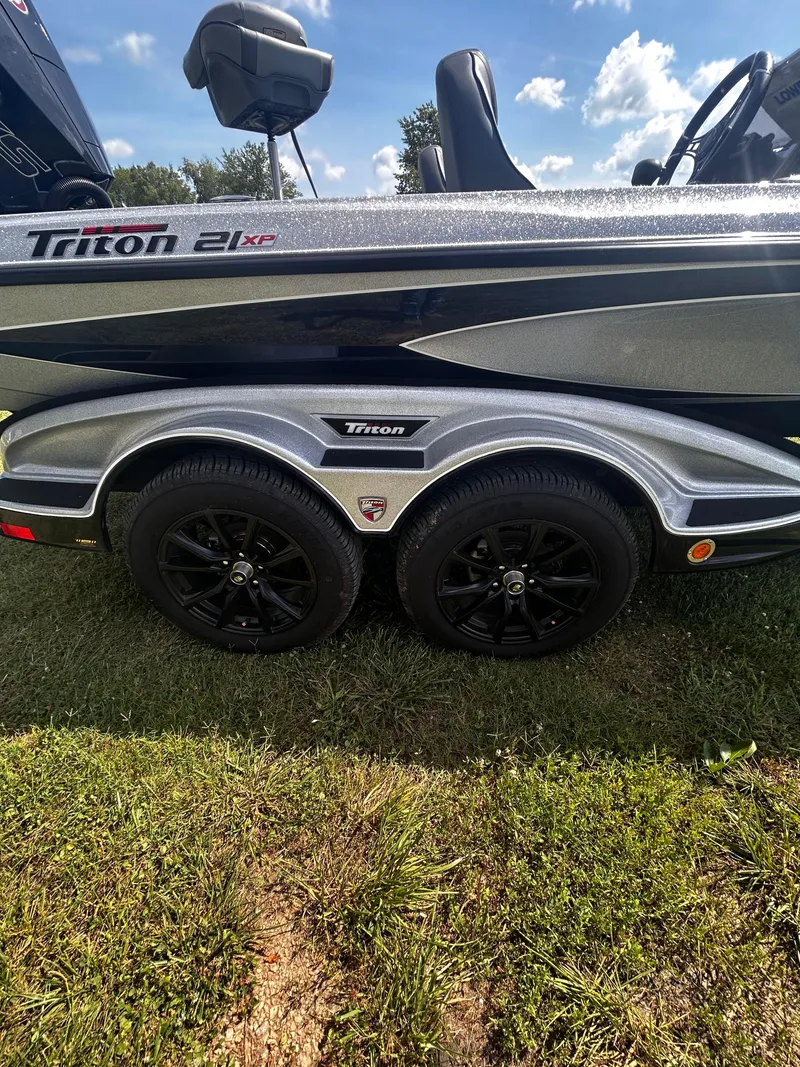 Slide: The Image of 2024 Triton 21XP boat trailer with black wheels on grass under a blue sky. - 21