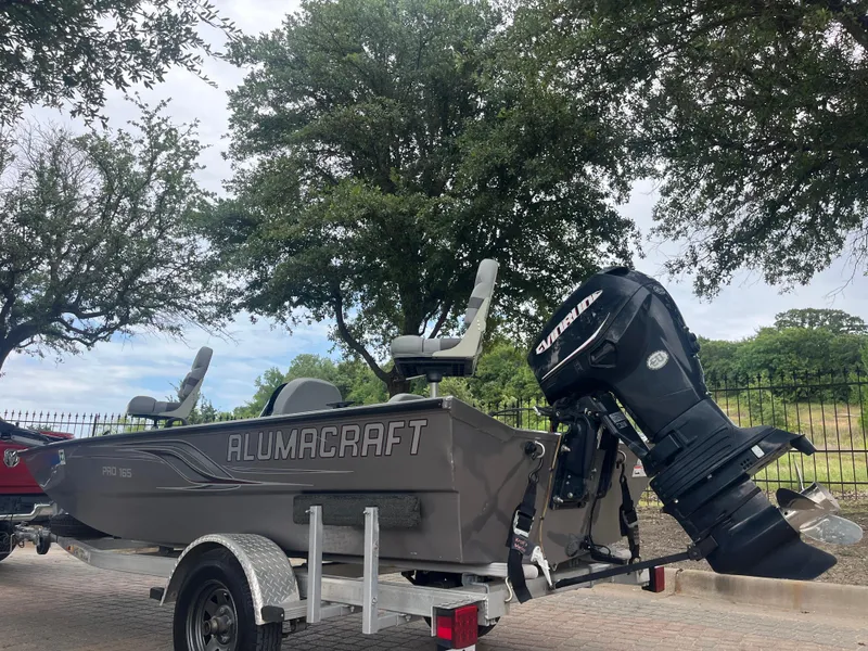Slide: The Image of 2012 Alumacraft Pro 165 boat on trailer with outboard motor, parked near trees. - 7