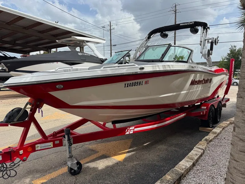Slide: The Image of 2018 MasterCraft XT23 boat on a red trailer, parked outdoors. - 2