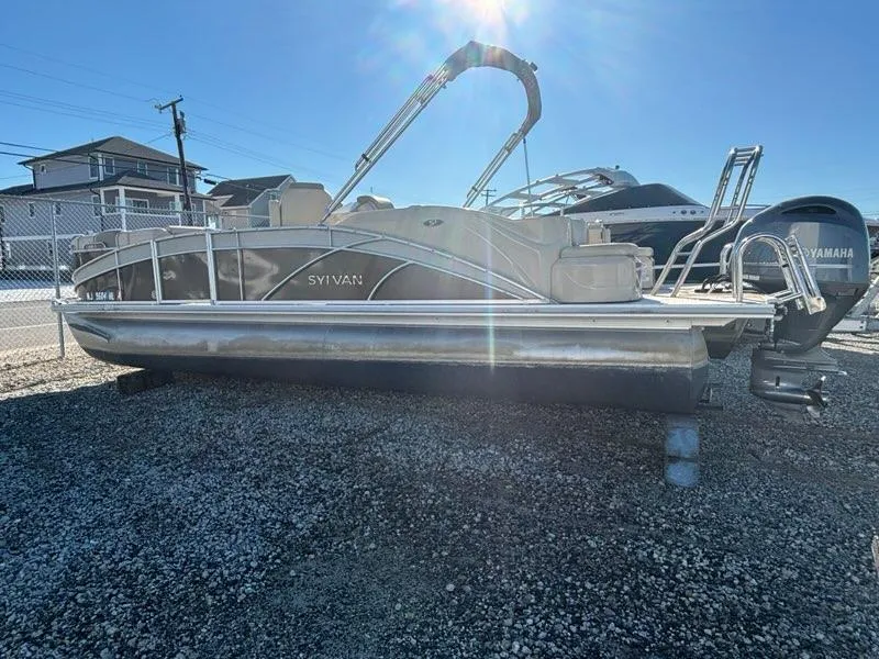 Slide: The Image of 2018 Sylvan S3 pontoon boat parked on gravel lot under bright sunlight. - 0