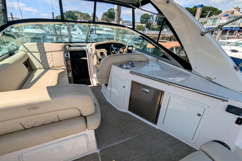 Slide: The Image of 2018 Regal 33 XO boat interior with beige seating, kitchenette, and steering area at a marina. - 12