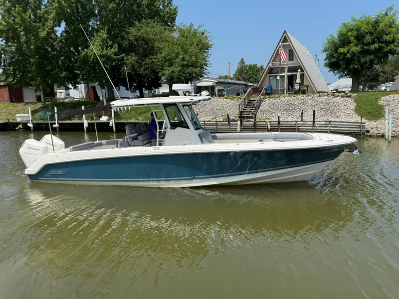 Slide: The Image of 2024 Boston Whaler 330 Outrage boat on calm water near a lakeside house. - 4