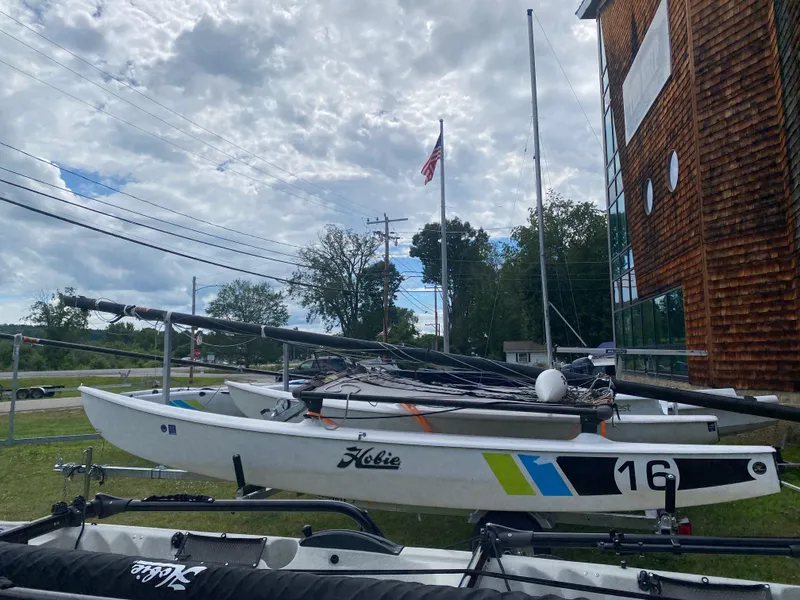 The Image of 2017 Hobie 16 catamaran on trailer, parked outdoors near wooden building and American flag. - 0