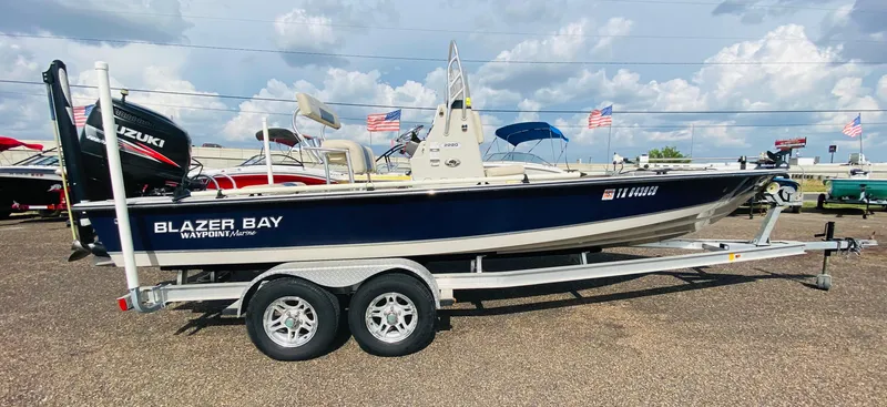 Slide: The Image of 2014 Blazer Bay 2200 Professional boat on trailer, displayed outdoors with flags in background. - 2