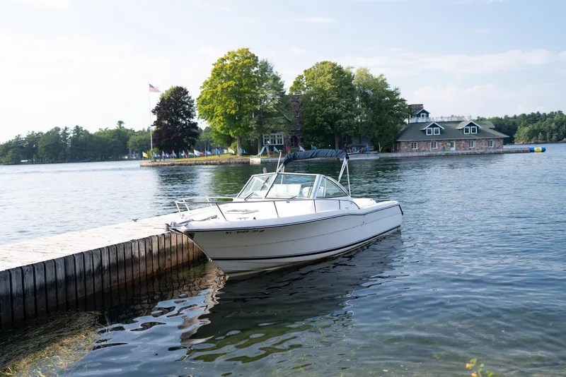 Slide: The Image of 2001 Pursuit 2460 Denali boat docked by a scenic lakeside with trees and buildings. - 1