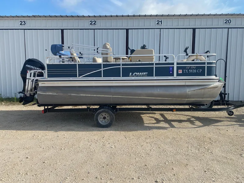 Slide: The Image of 2014 Lowe SF194 Sport Fish pontoon boat on trailer, parked near storage units. - 0