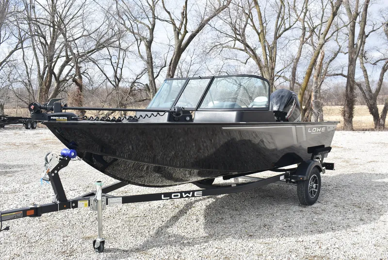 Slide: The Image of 2025 Lowe FM 1675 WT fishing boat on trailer, parked outdoors with trees in background. - 3