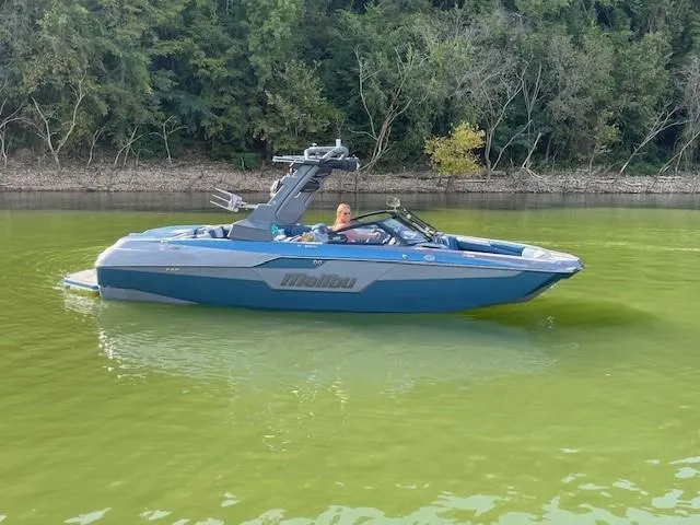 Slide: The Image of 2023 Malibu M240 boat on a calm green lake with forested shoreline. - 2
