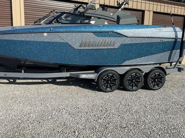 Slide: The Image of 2023 Malibu M240 boat on trailer, blue exterior, parked on gravel. - 3