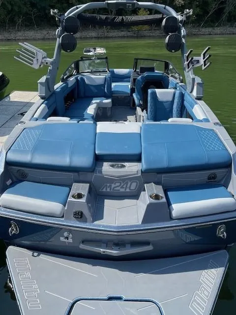 Slide: The Image of 2023 Malibu M240 boat with blue seating, spacious deck, and modern design on a calm lake. - 7