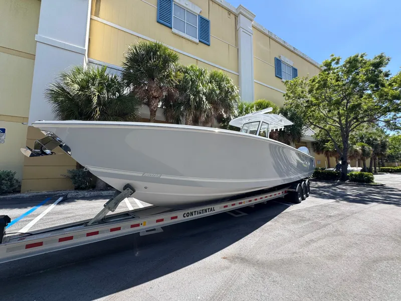 Slide: The Image of 2025 Jupiter 380LS boat on trailer, parked near a building with palm trees. - 2