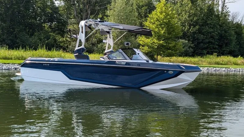 Slide: The Image of 2025 Nautique Super Air Nautique GS22 boat on a calm lake with lush greenery. - 3