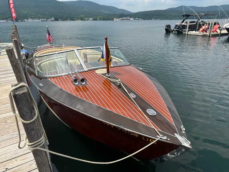 Slide: The Image of 1999 Hacker-Craft Custom Sport boat docked on a serene lake with scenic mountain backdrop. - 6