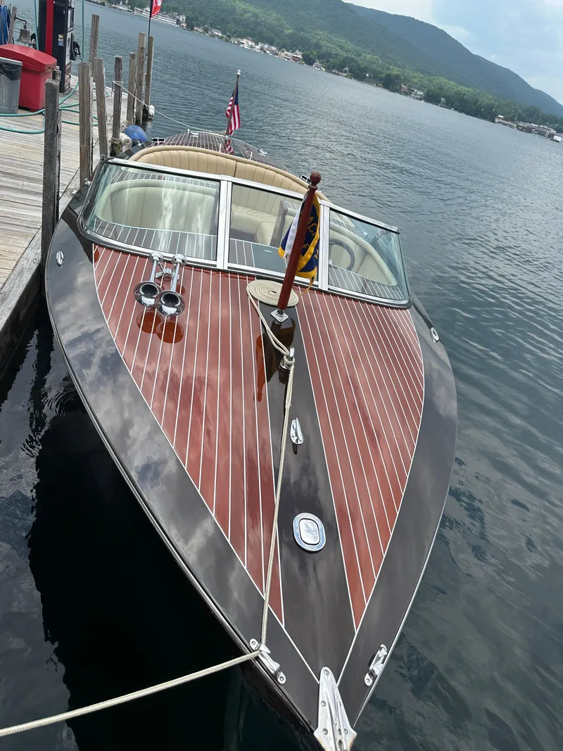 Slide: The Image of 1999 Hacker-Craft Custom Sport boat docked on a serene lake with mountains in the background. - 7