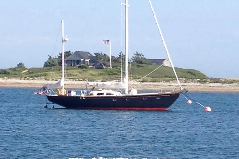 Slide: The Image of Sailboat anchored near shore, Fastnet Lecomte 1972 model, with house and American flag in background. - 0