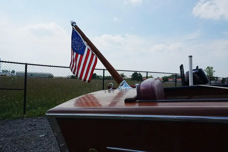 Slide: The Image of 1956 Chris-Craft Continental boat with American flag, parked near a fence under a cloudy sky. - 16