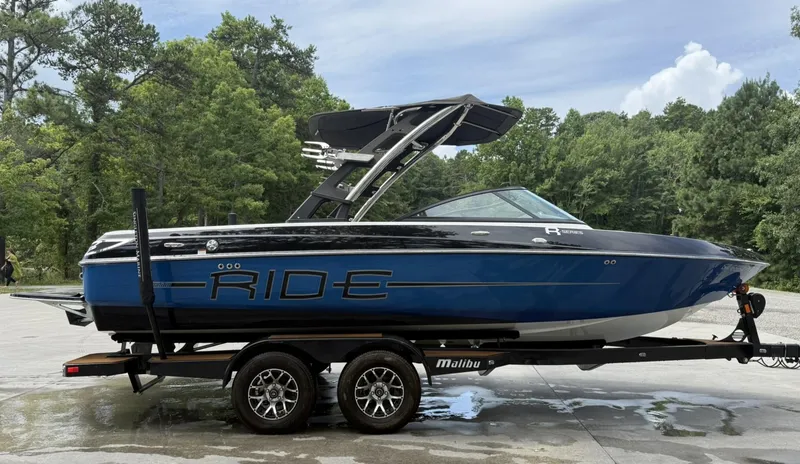 Slide: The Image of 2012 Malibu 21 Ride boat on trailer, parked outdoors with trees in background. - 3