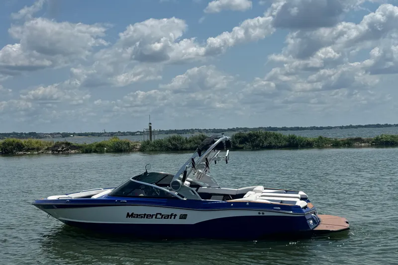 The Image of 2021 MasterCraft XT23 boat on a calm lake under a partly cloudy sky. - 0