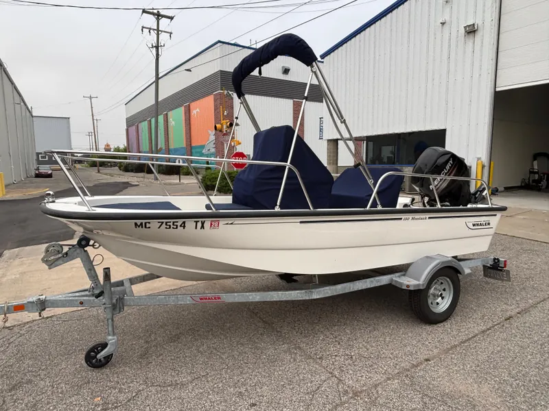 Slide: The Image of 2016 Boston Whaler 150 Montauk boat on trailer, parked near industrial buildings. - 2
