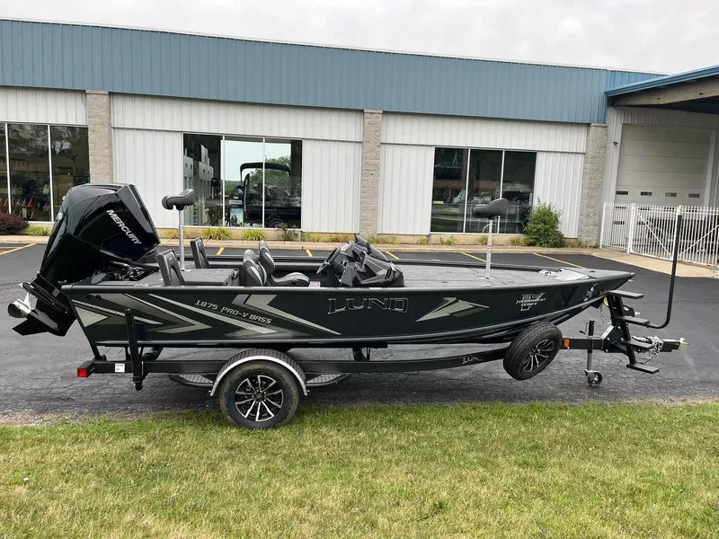 The Image of 2026 Lund 1875 Pro-V Bass XS boat on trailer, parked outside a building. - 0