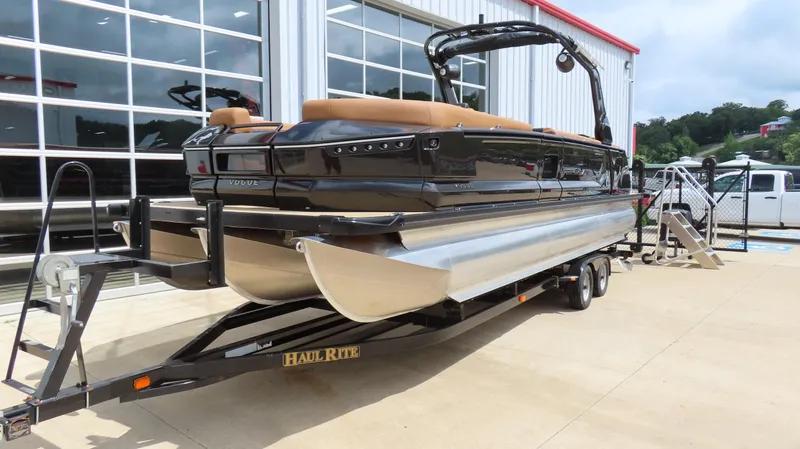 Slide: The Image of 2025 Princecraft Vogue 27 RL pontoon boat on trailer, parked outside a modern building. - 2