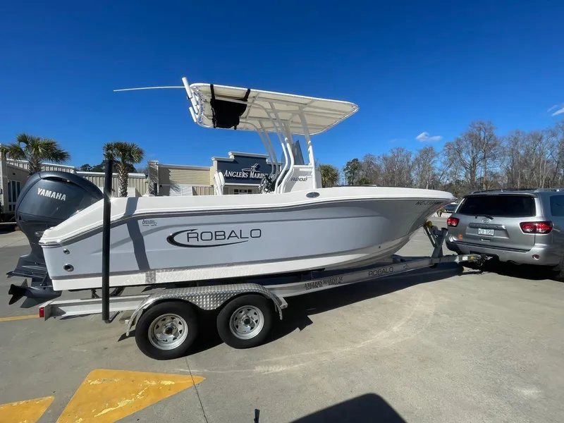 Slide: The Image of 2023 Robalo R202 Explorer boat on trailer, parked beside SUV, clear blue sky. - 0