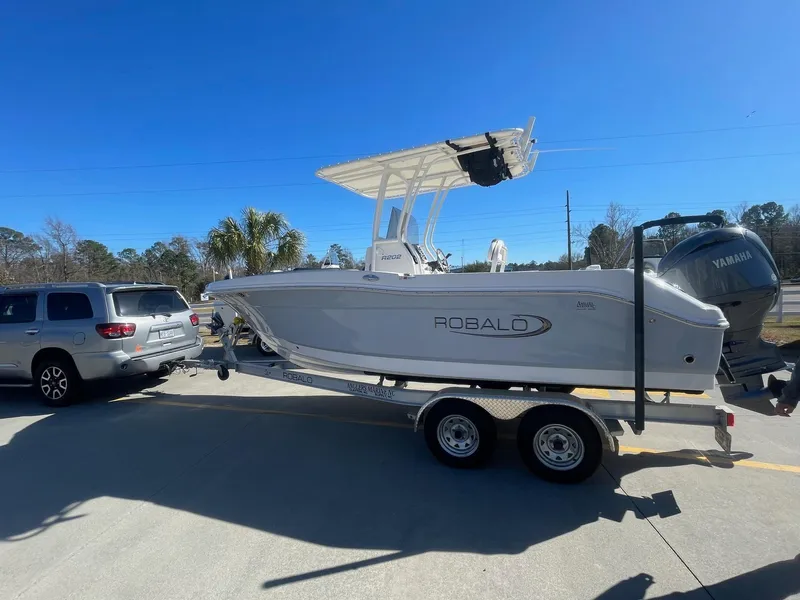 Slide: The Image of 2023 Robalo R202 Explorer boat on trailer, parked beside SUV, under clear blue sky. - 2