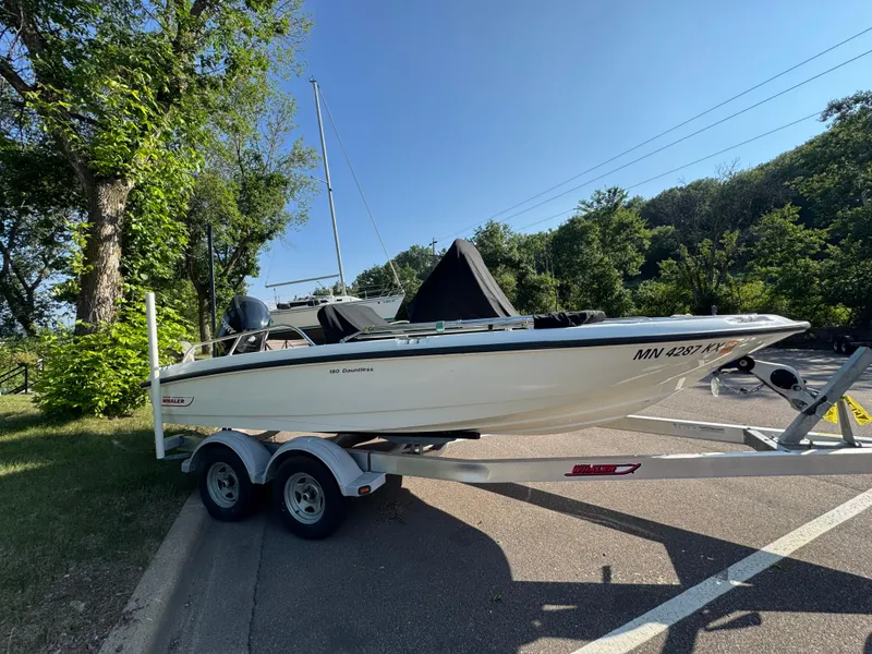 Slide: The Image of 2011 Boston Whaler 180 Dauntless boat on trailer, parked near trees under clear sky. - 3