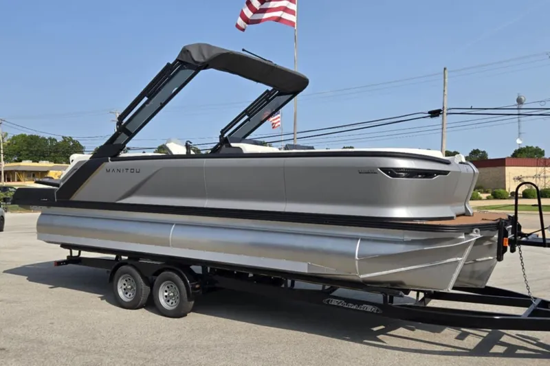Slide: The Image of 2025 Manitou Explore pontoon boat on trailer, parked outdoors with American flag. - 1