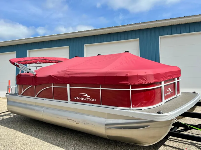 Slide: The Image of 2023 Bennington 20 SVSR pontoon boat with red cover, parked outside a blue building. - 2