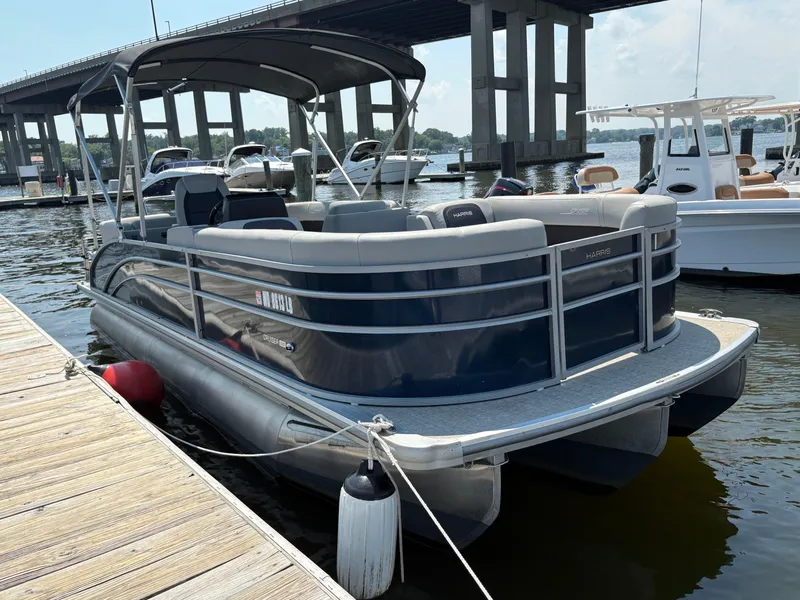 Slide: The Image of 2024 Harris Cruiser 230 pontoon boat docked near a bridge on a sunny day. - 0